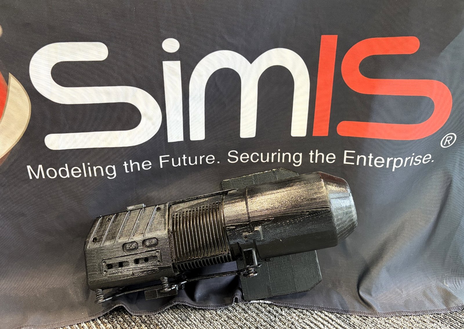 Black FDM 3D-printed assembly with visible layer lines, photographed in front of a SimIS banner.