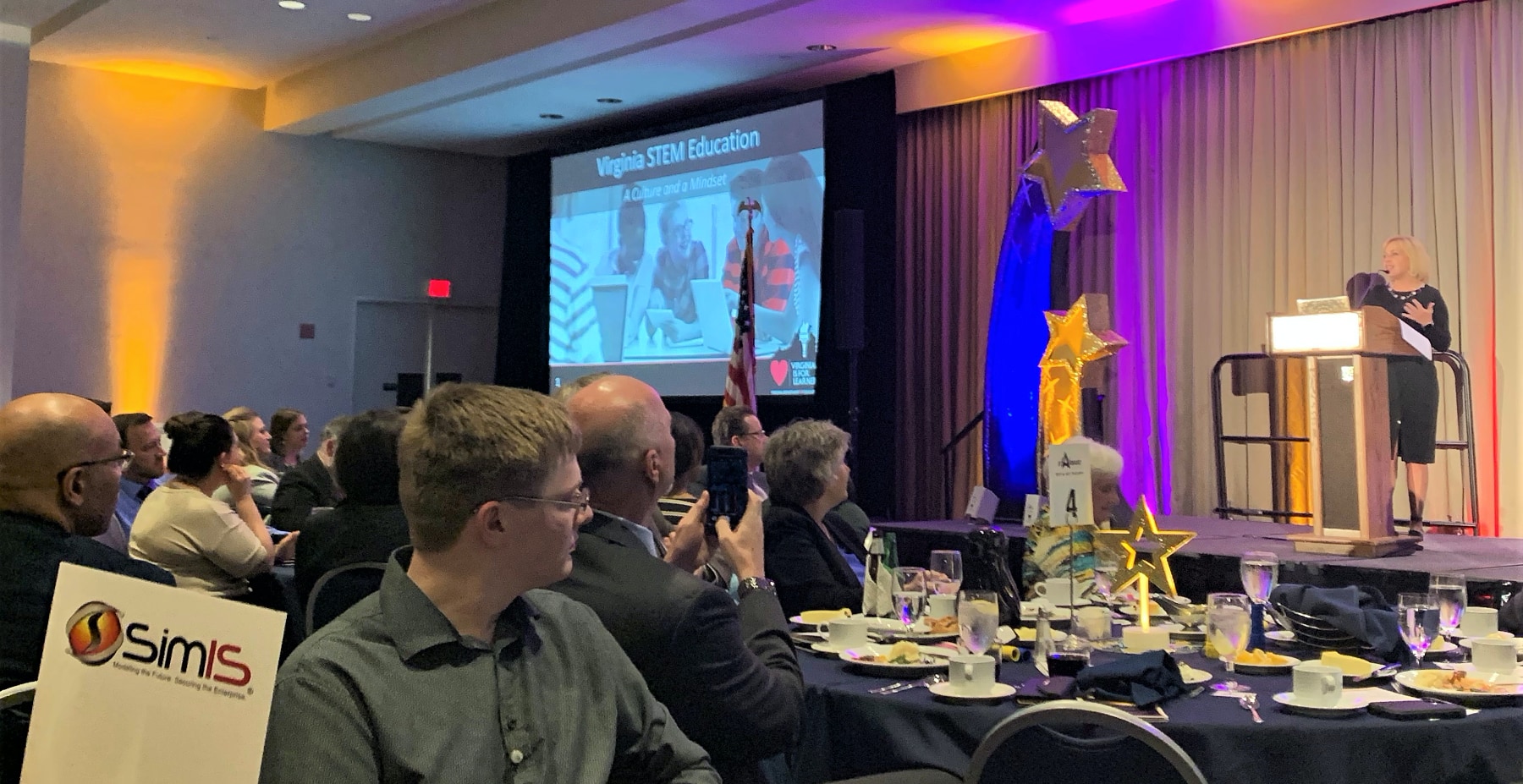 SimIS attendees at their table, with keynote speaker Dr. Tina Manglicmot presenting on Virginia STEM Education at the Launching Futures Event.