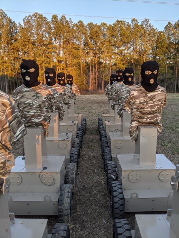 Two rows of Human Type Target robots wearing camouflage shirts and balaclava-style heads, lined up outdoors facing each other.